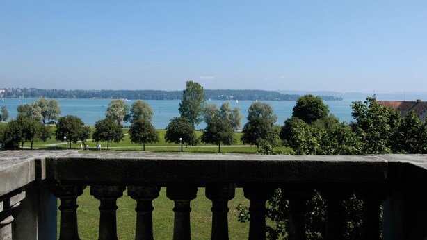 Une vue depuis un balcon donnant sur une étendue d’eau, avec des arbres et une silhouette de ville en arrière-plan.