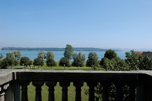 Une vue depuis un balcon donnant sur une étendue d’eau, avec des arbres et une silhouette de ville en arrière-plan.