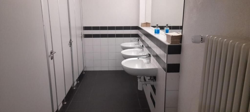 A narrow bathroom with three sinks, a mirror, and a radiator.
