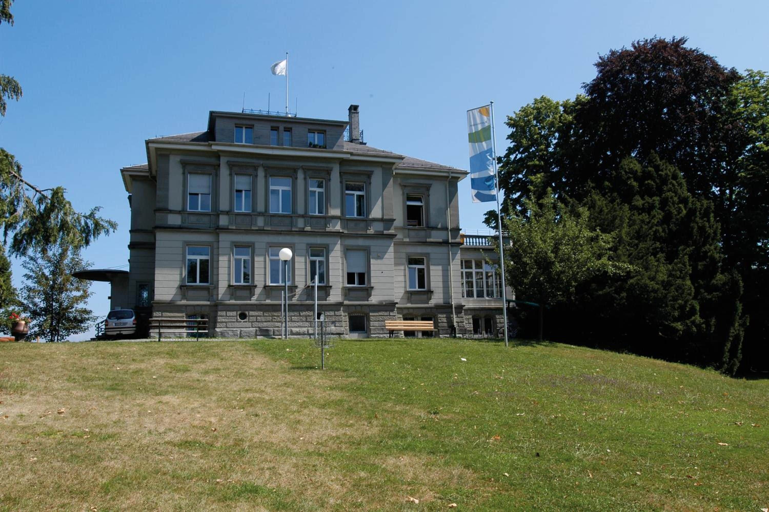Un edificio grande de varios pisos con fachada simétrica, un asta de bandera en el techo y un área verde en primer plano.