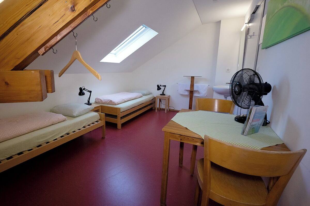 Une petite pièce compacte équipée de deux lits superposés, d’un bureau, d’une chaise, d’un ventilateur et d’une lucarne.