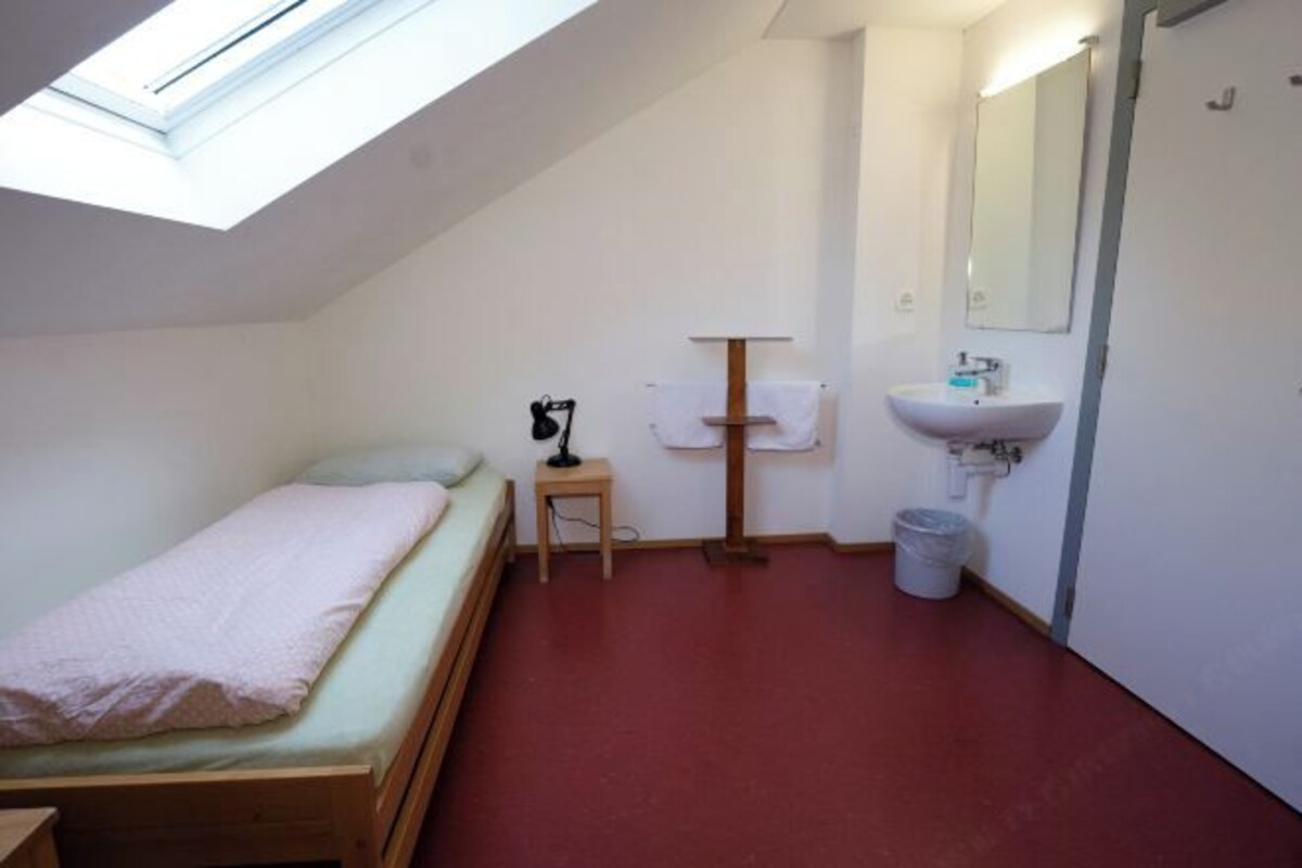 A small, single-bed room with a skylight, a bedside table with a lamp, a wall-mounted sink, and a trash bin.