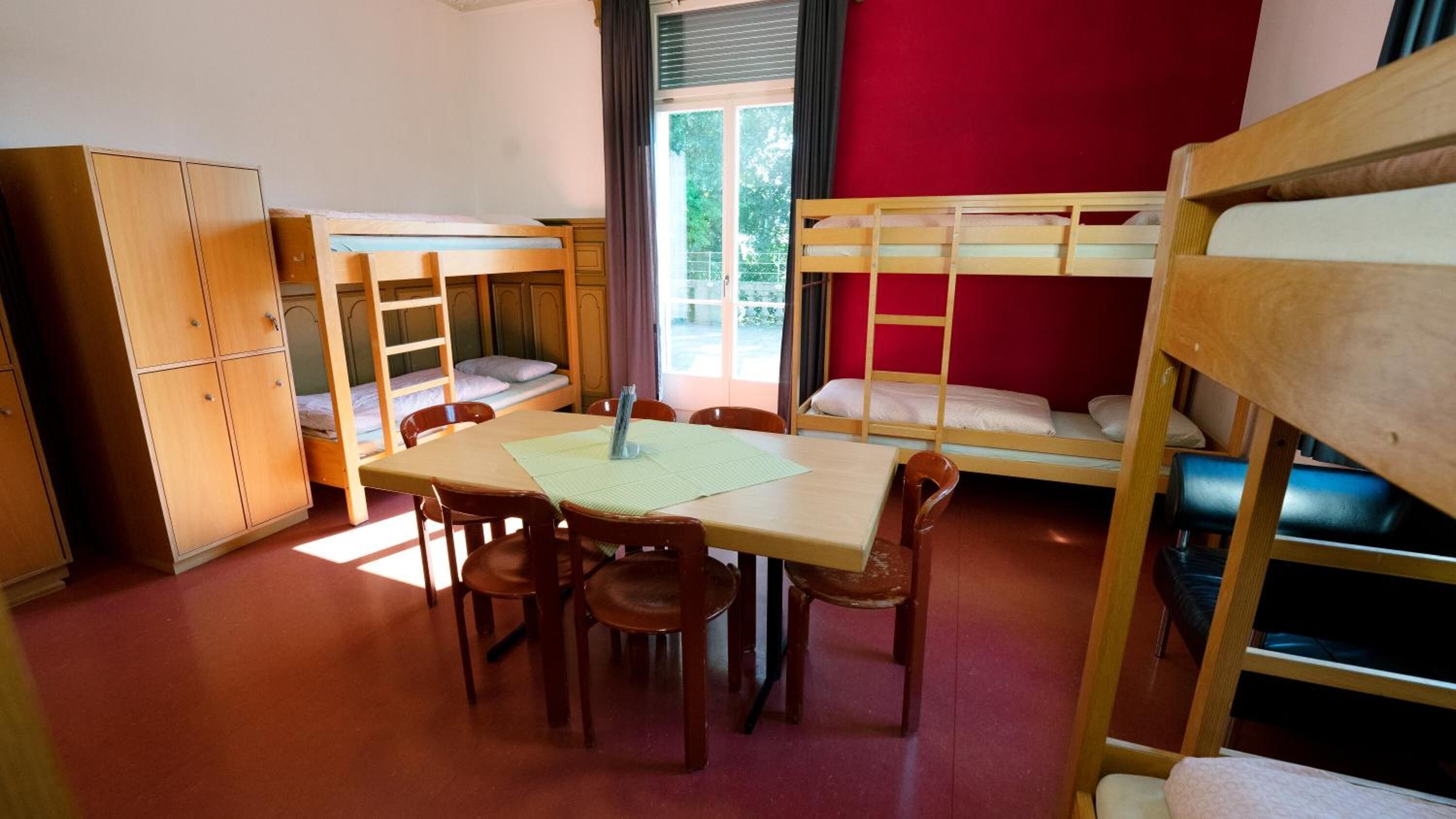 A dormitory room with bunk beds, a wooden desk, chairs, and a wardrobe.