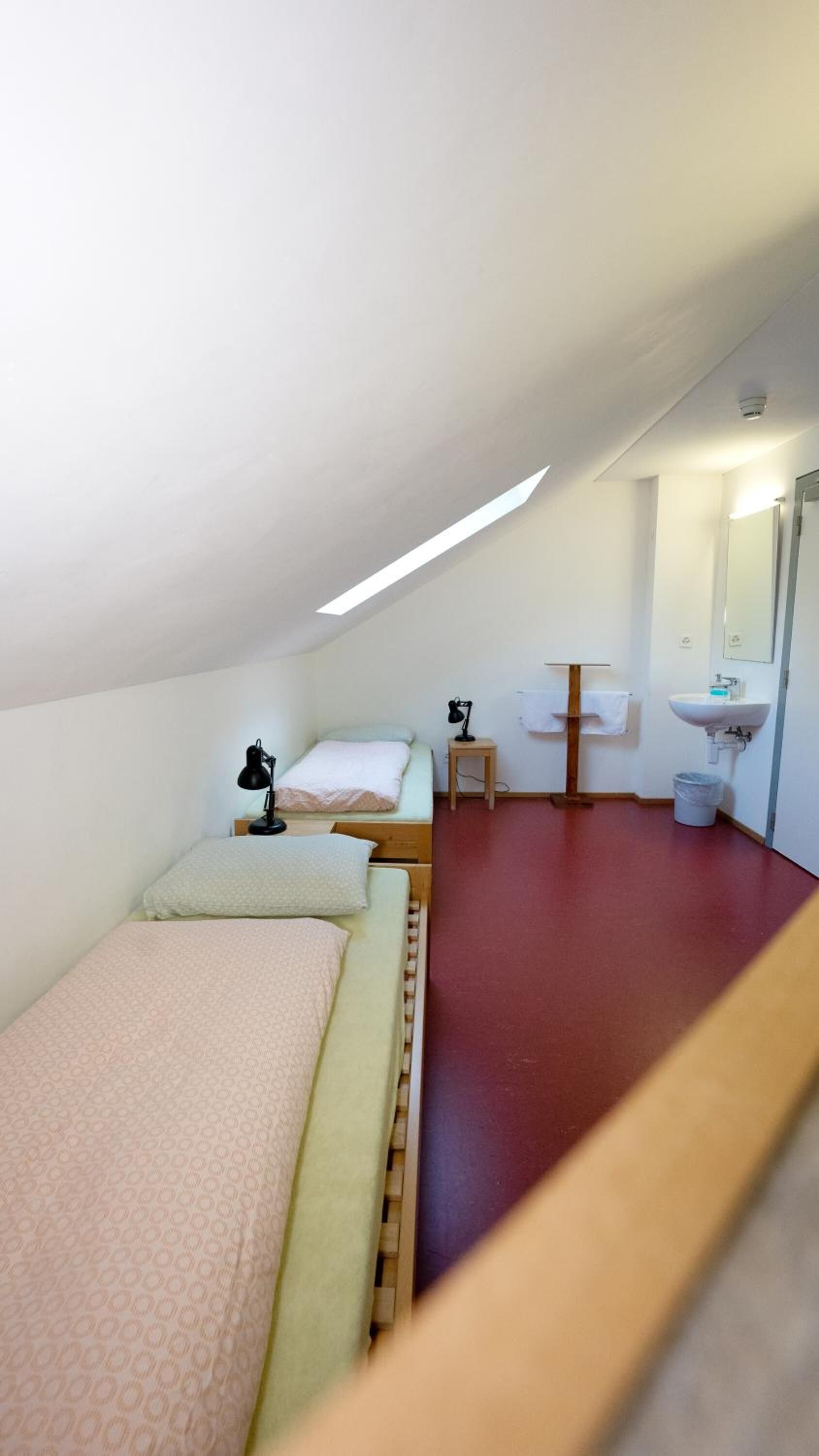 Une petite pièce, de taille modeste, avec un plafond en pente, un lit simple, une table de chevet avec une lampe, un petit tabouret, un miroir et un lavabo.