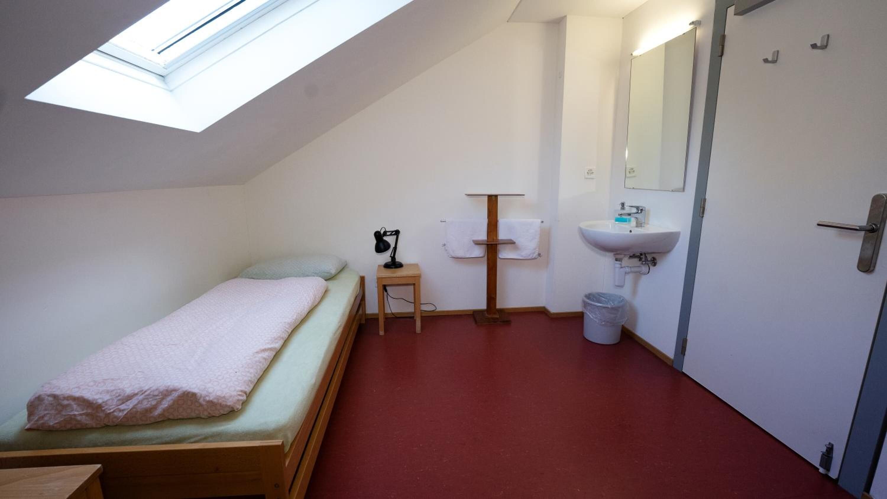 Une petite pièce simple, avec un lit, un bureau, un miroir, un lavabo et une poubelle.