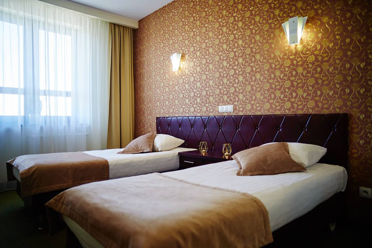 Ein Hotelzimmer mit zwei Betten, einem großen Fenster mit durchscheinenden Vorhängen und einer Wand mit Musterpapier.