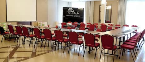 A conference room with a large table, red chairs, a projector screen, and a banner with hotel branding.