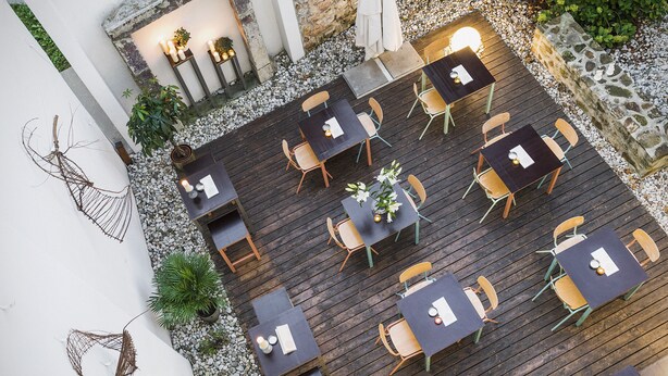 Un patio al aire libre con varias mesas y sillas, rodeado por un muro de piedra y una pared blanca con detalles decorativos.