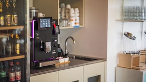 En kaffemaskine med digitalt display, en vask og hylder med diverse genstande, herunder flasker og glas.
