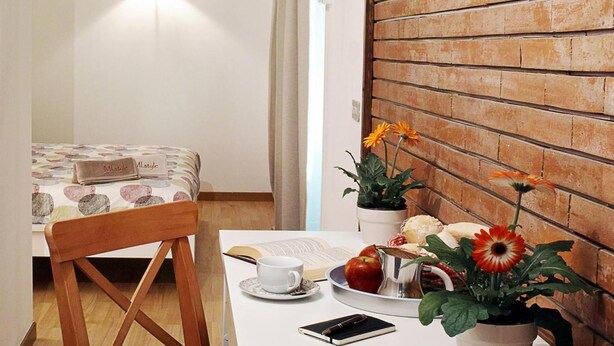 A small room with a bed, a wooden chair, a table with a book, a cup, a plate with food, and a potted plant. - MI.style (Milan)