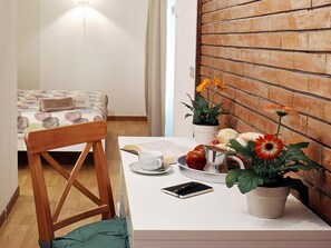 Una habitación pequeña con una cama, una silla de madera, una mesa con un libro, una taza, un plato con comida y una planta en maceta.