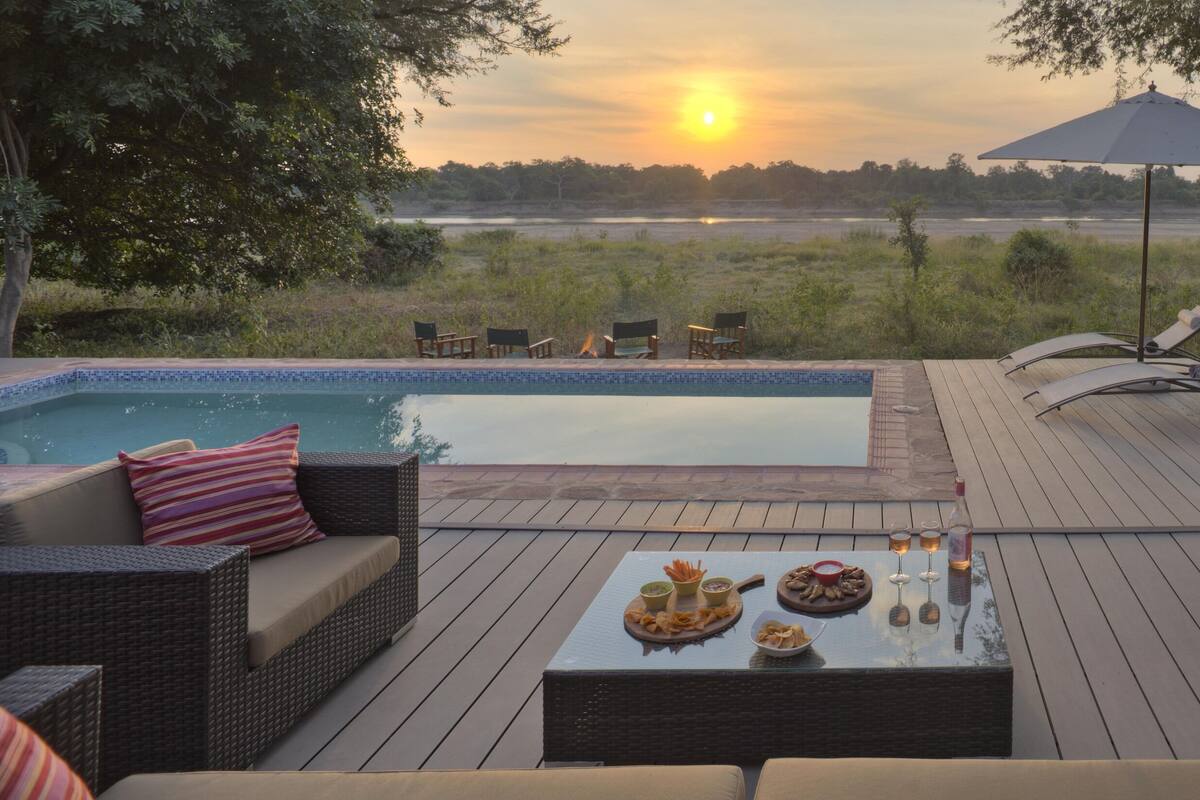 Ein Poolbereich mit Korbmöbeln, einem Tisch mit Snacks und Getränken und Blick auf den Sonnenuntergang über dem Wasser.