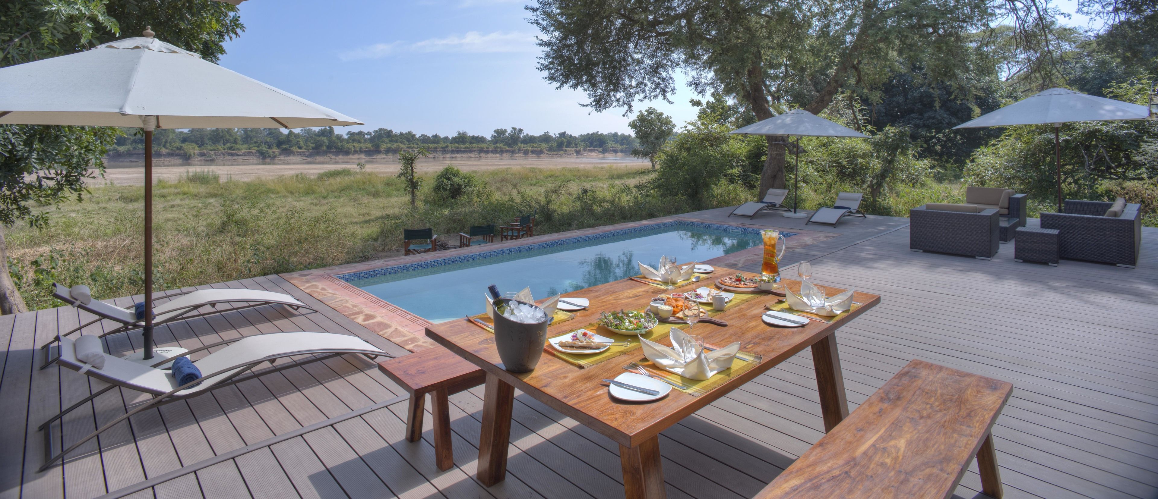 Un área de comedor al aire libre con piscina, terraza de madera, sillones de descanso y sombrillas.