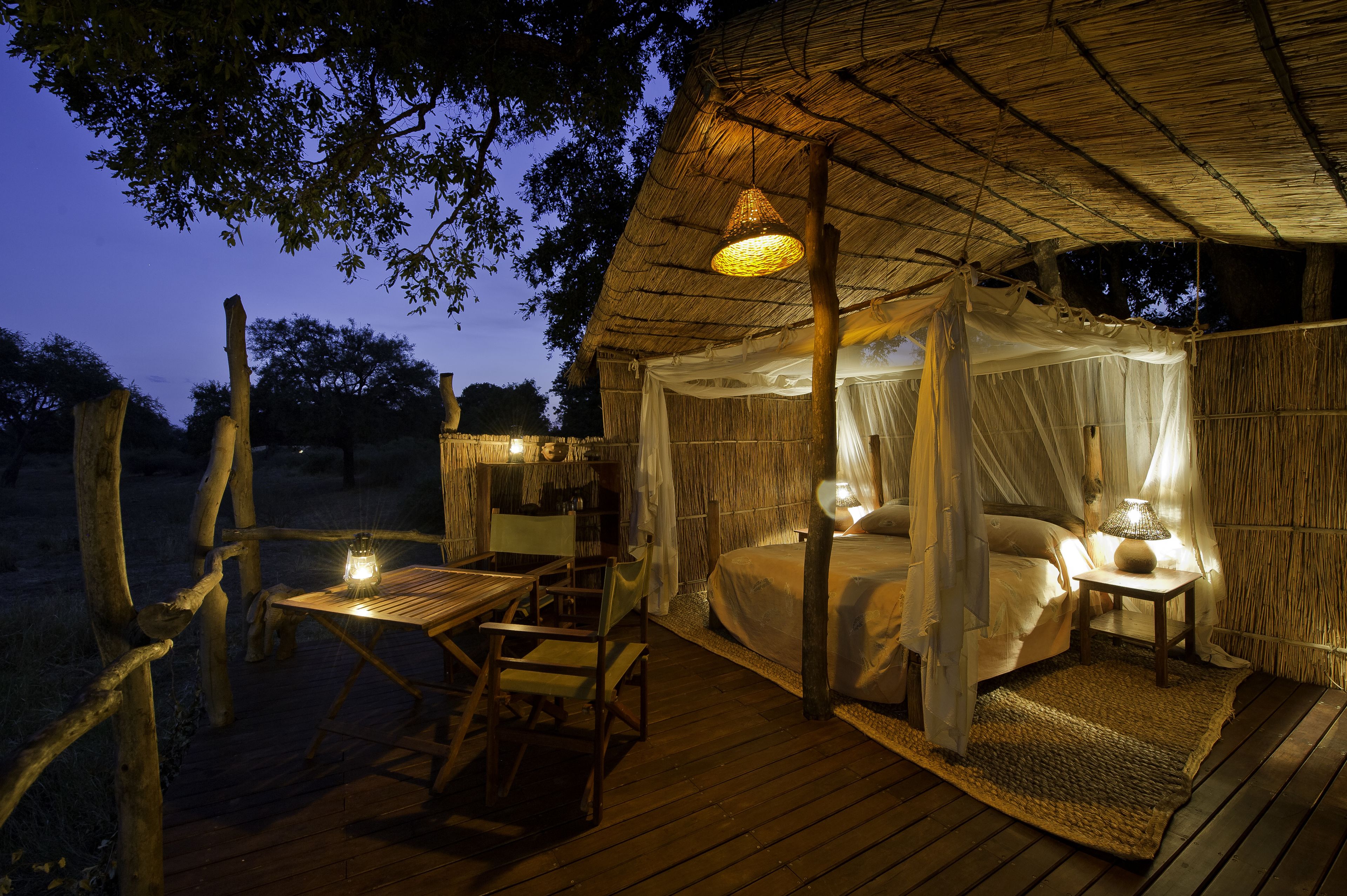 Dormitorio al aire libre con techo de paja, terraza de madera, mesa con sillas y una lámpara de noche.