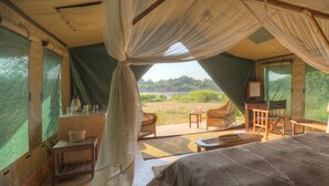 A tented room with a bed, a table, and chairs, and a view of a green landscape.