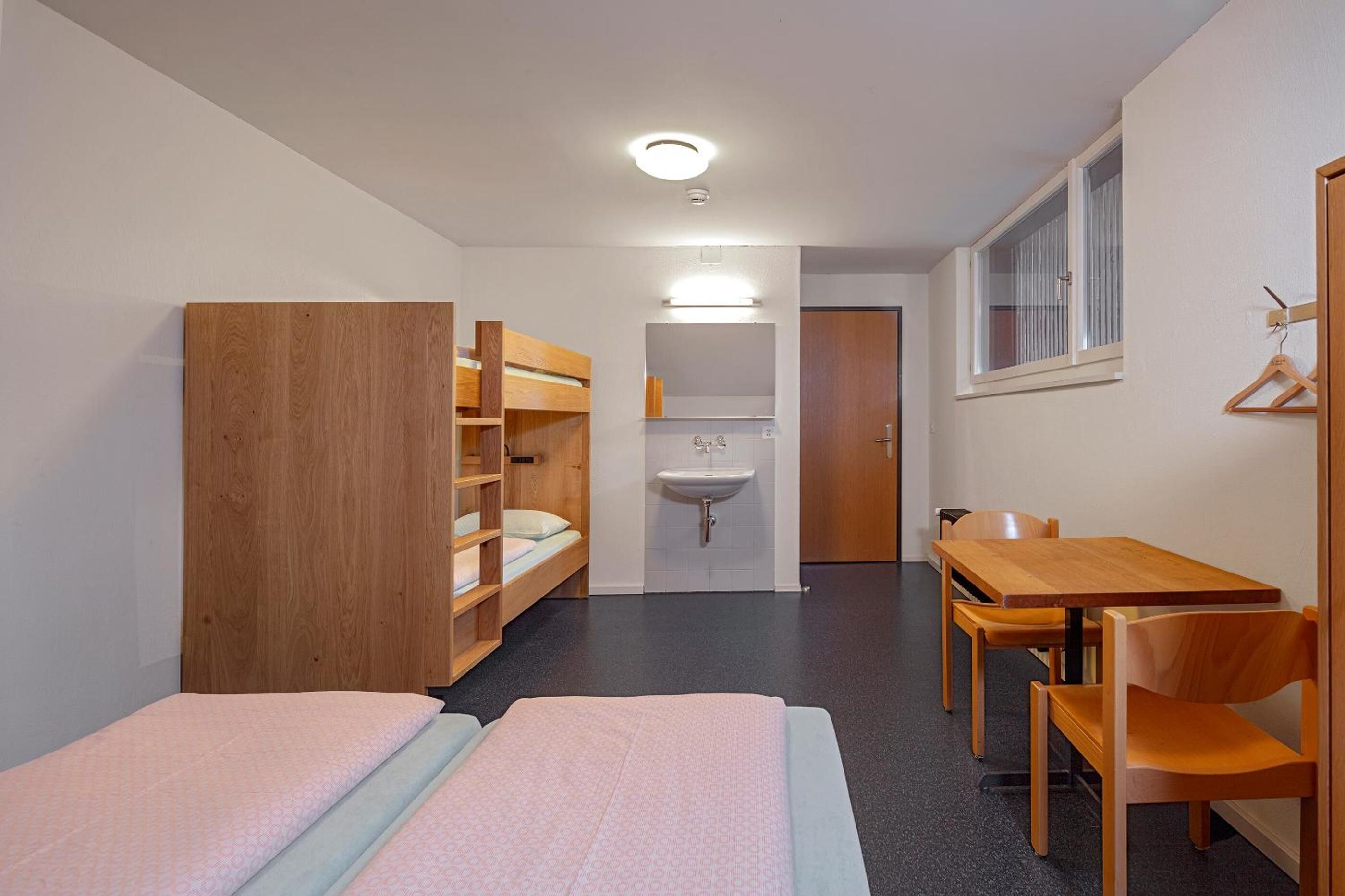 Habitación tipo dormitorio con dos camas, un armario de madera, un lavamanos, una mesa y sillas.