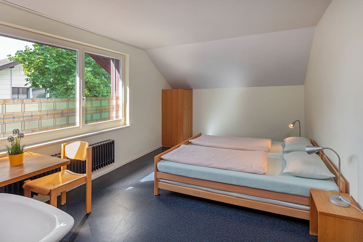 A bedroom with a bed, a desk with a chair, a radiator, and a window with a view of greenery.