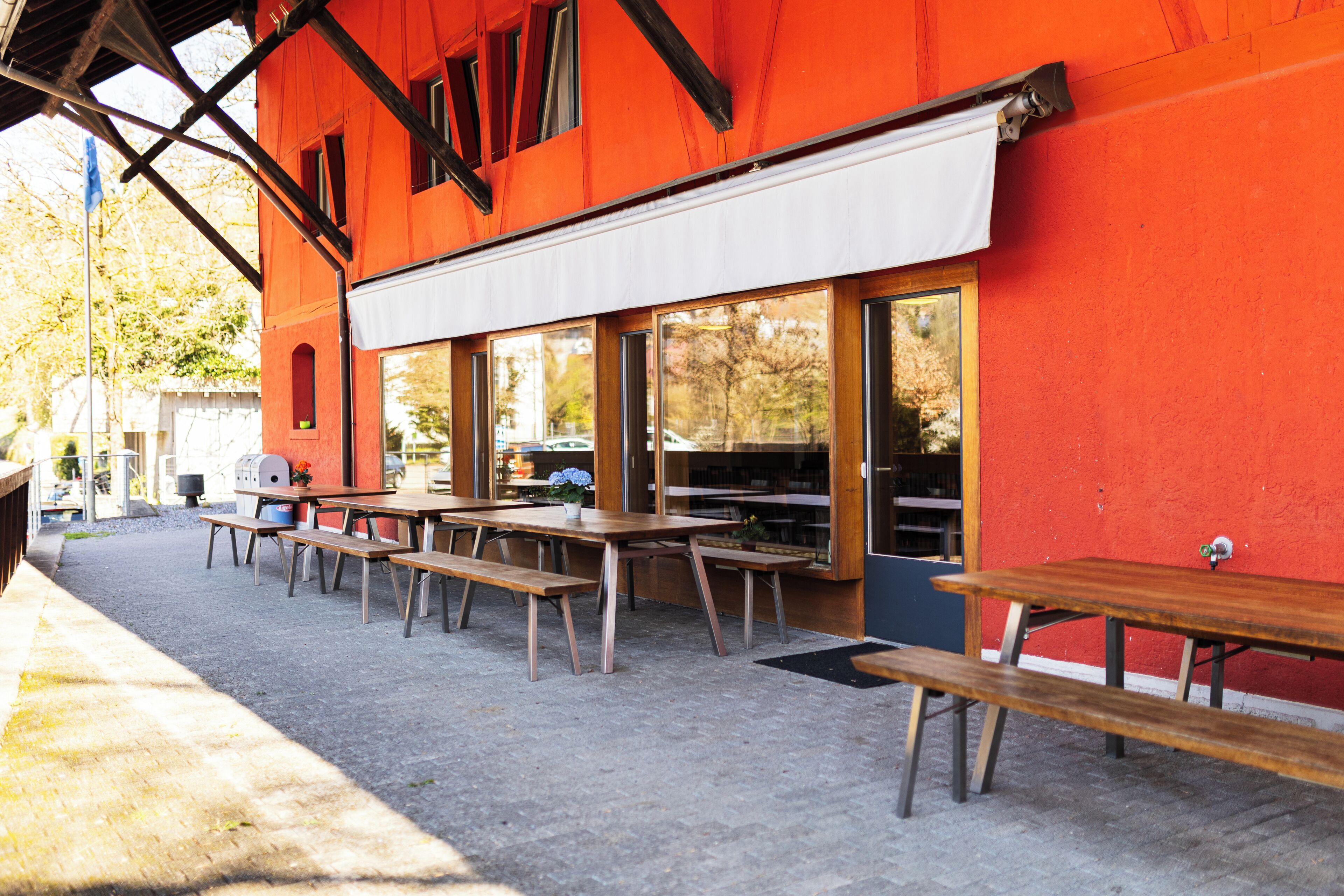 An outdoor seating area with wooden tables and benches, a covered patio, and a red building facade.