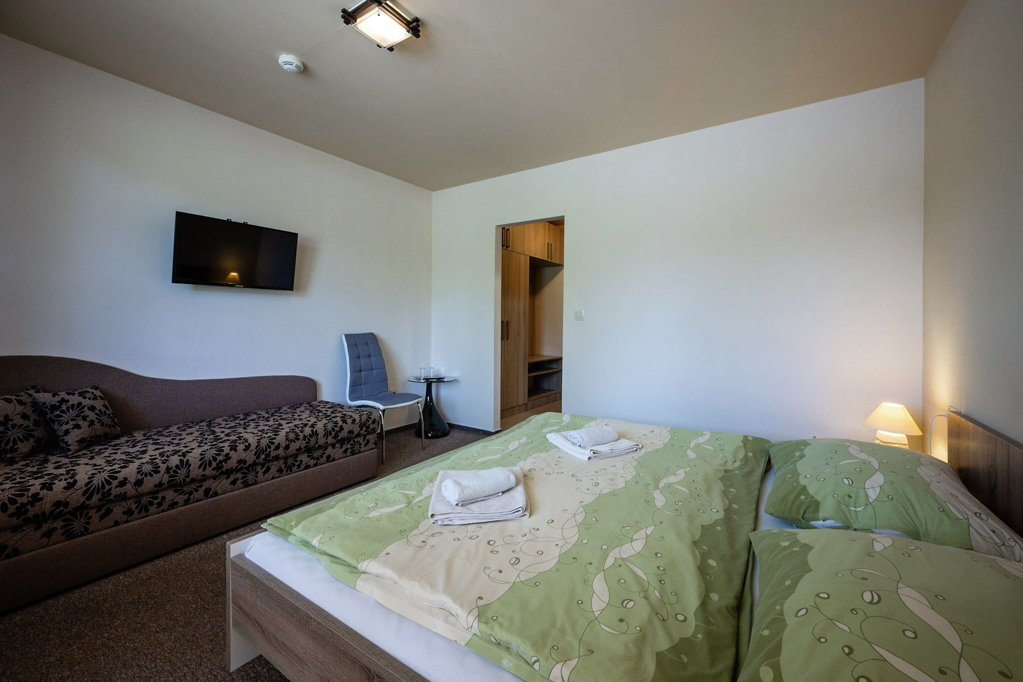 A hotel room with a sofa, a bed with green and beige bedding, a flat-screen TV mounted on the wall, and a small table with a chair.