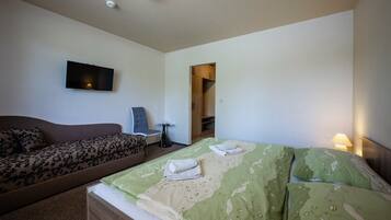 A hotel room with a sofa, a bed with green and beige bedding, a flat-screen TV mounted on the wall, and a small table with a chair.