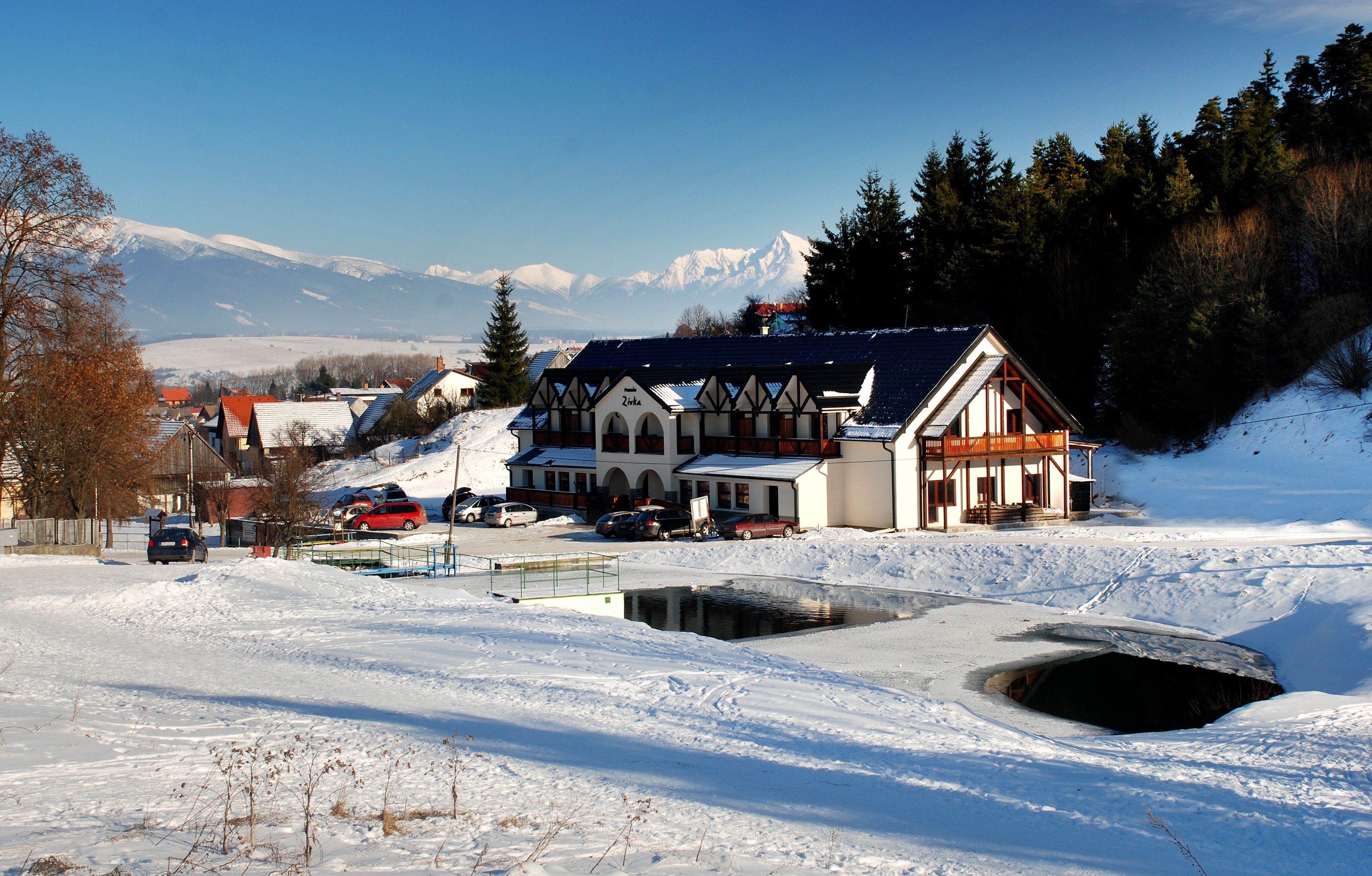 Un bâtiment de style chalet traditionnel, entouré d’un paysage enneigé, avec un petit étang et une chaîne de montagnes en arrière-plan.