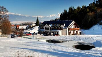 Un bâtiment de style chalet traditionnel, entouré d’un paysage enneigé, avec un petit étang et une chaîne de montagnes en arrière-plan.