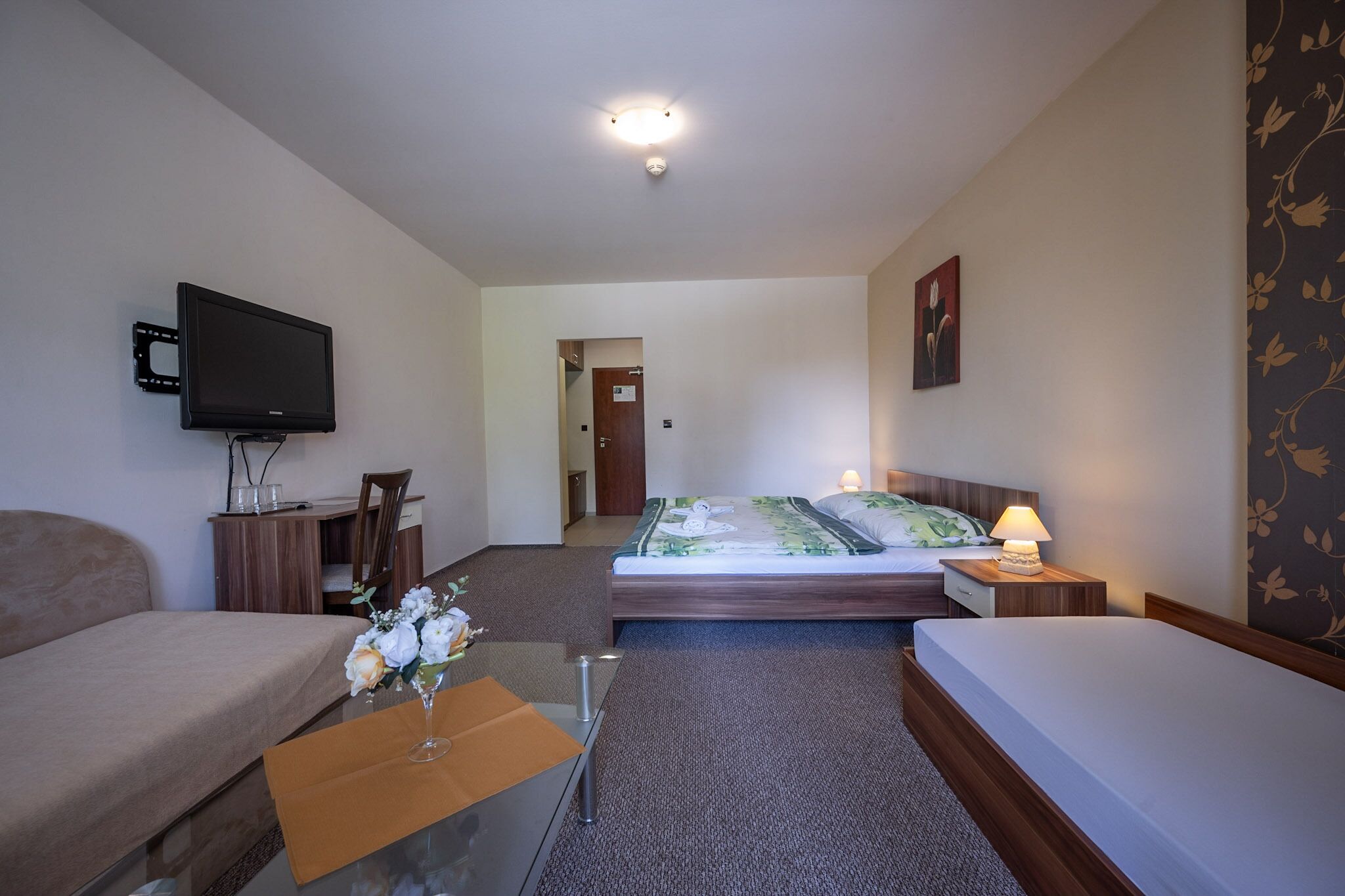 Une chambre d’hôtel avec un lit, un canapé, une télévision, une petite table ornée d’un vase de fleurs et une table de chevet avec une lampe.