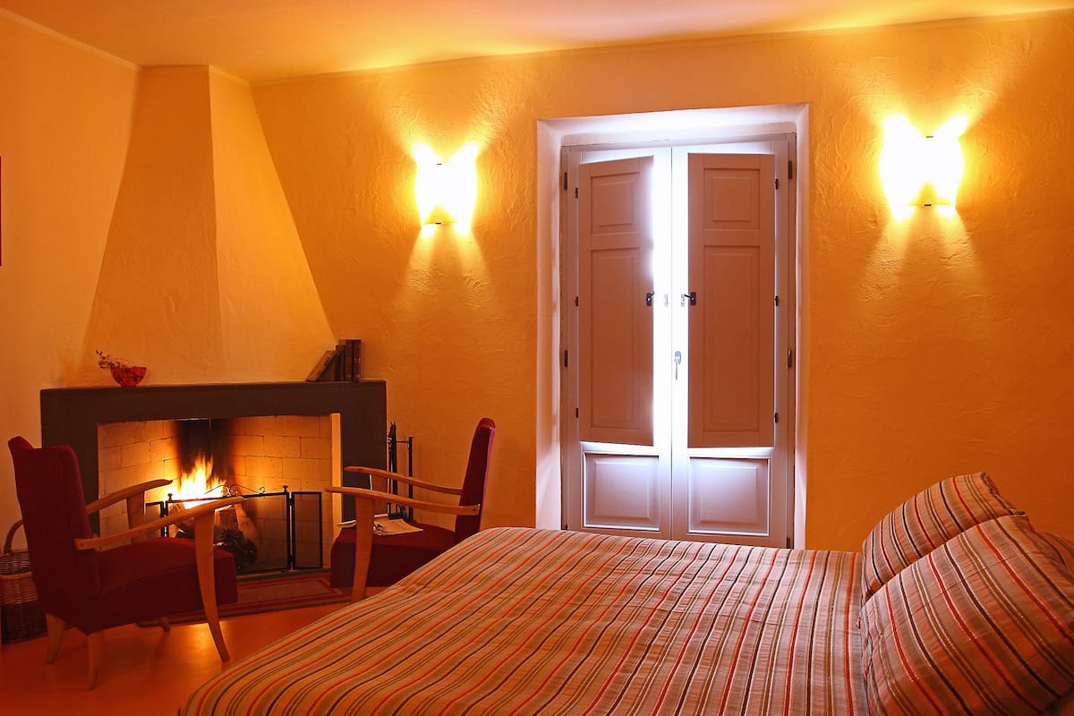 A bedroom with a bed, a chair, a fireplace, and a door with light fixtures.
