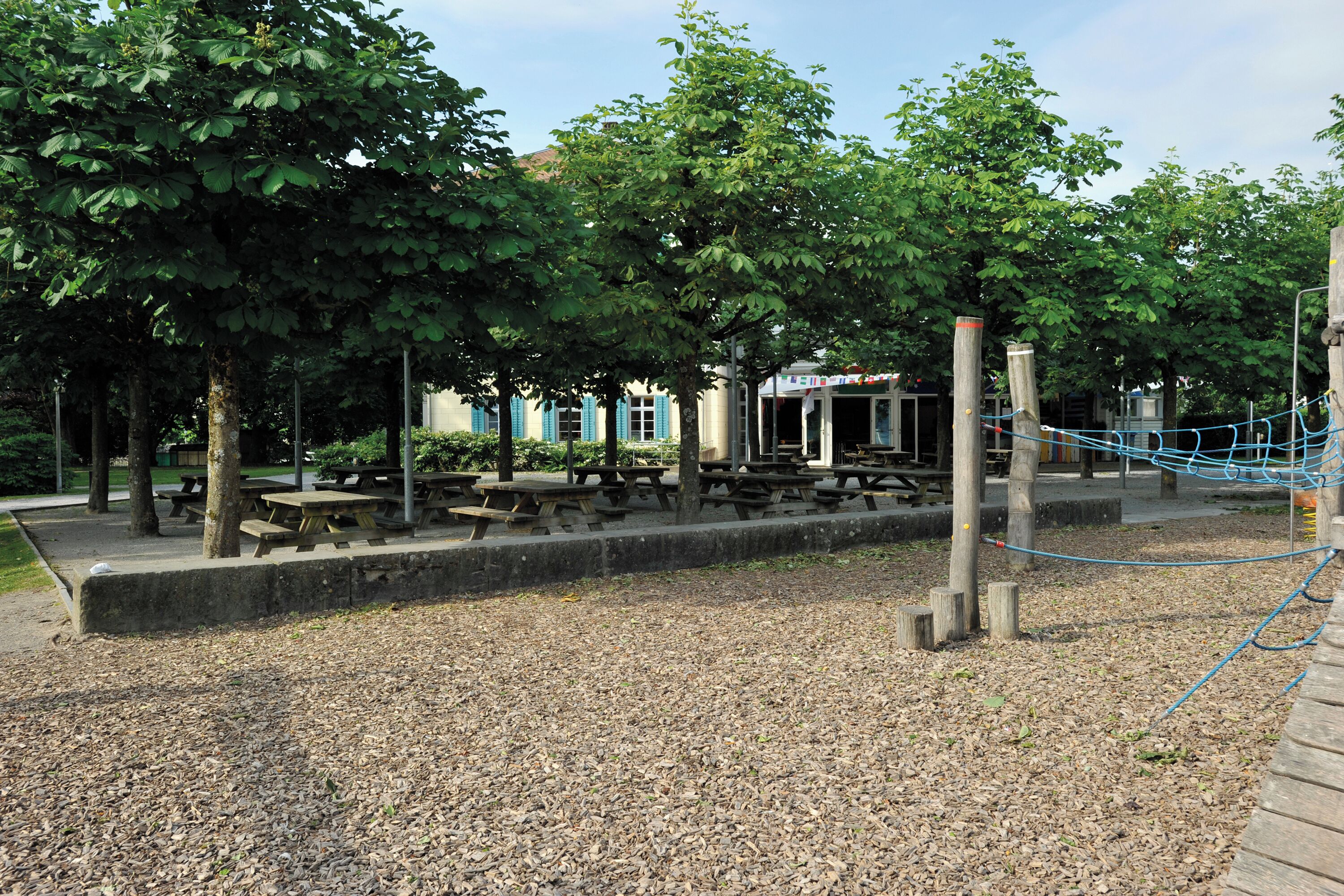 A park with picnic tables, a playground, and a building with blue shutters.