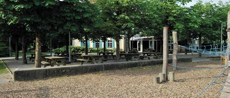 A park with picnic tables, a playground, and a building with blue shutters.