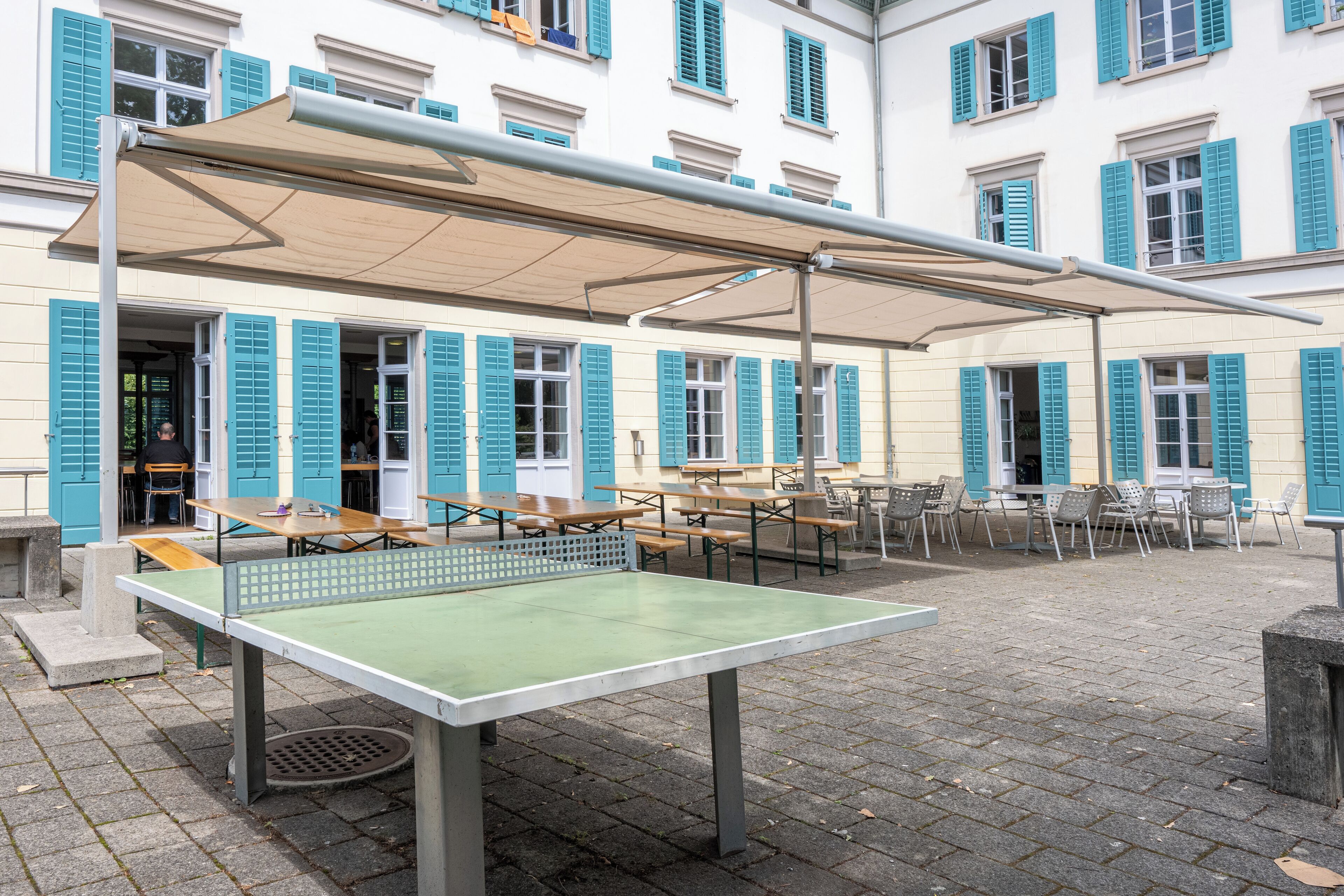 Una mesa de tenis de mesa al aire libre con toldo, rodeada de asientos exteriores y un edificio con contraventanas azules al fondo.
