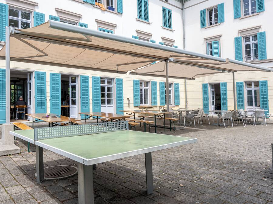 An outdoor table tennis table with a canopy, surrounded by outdoor seating and a building with blue shutters in the background.