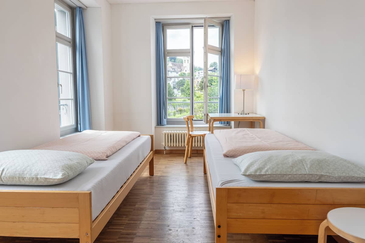 Ein Zimmer mit zwei Holzbetten, einem Sessel, einem kleinen Tisch und einem Fenster mit Vorhängen.