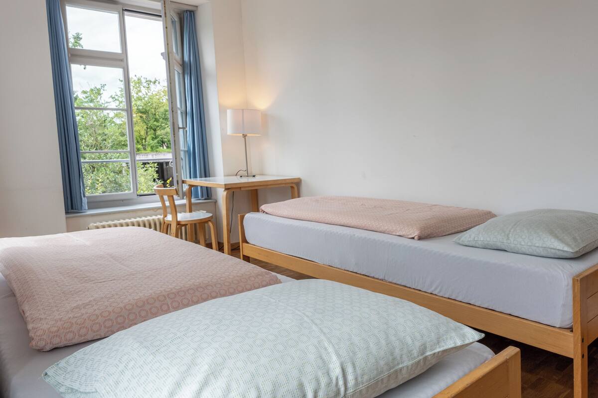 Ein Zimmer mit zwei Einzelbetten, einem kleinen Tisch, einem Stuhl und einem Fenster mit Vorhängen.
