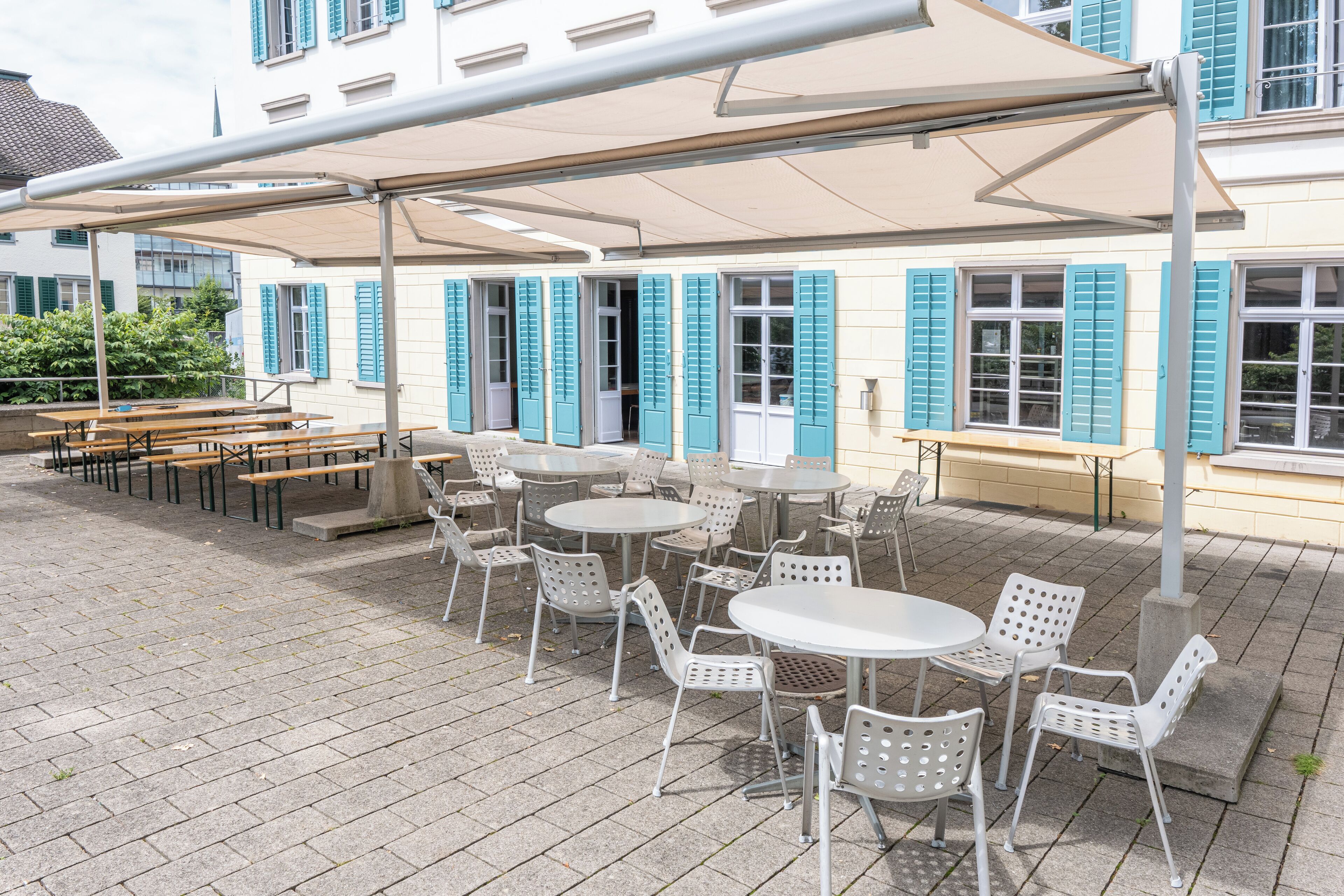 An outdoor patio with tables and chairs, a covered seating area, and a building with blue shutters in the background.