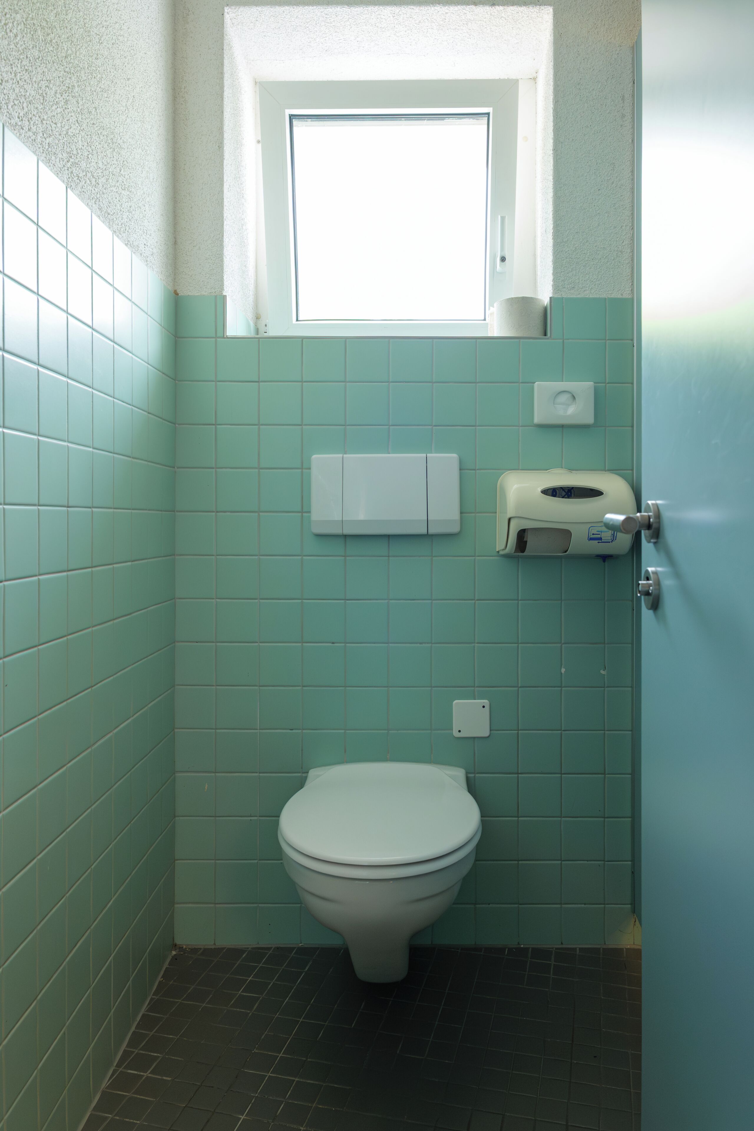 A bathroom with teal tiles, a toilet, a paper towel dispenser, and a window above the toilet.