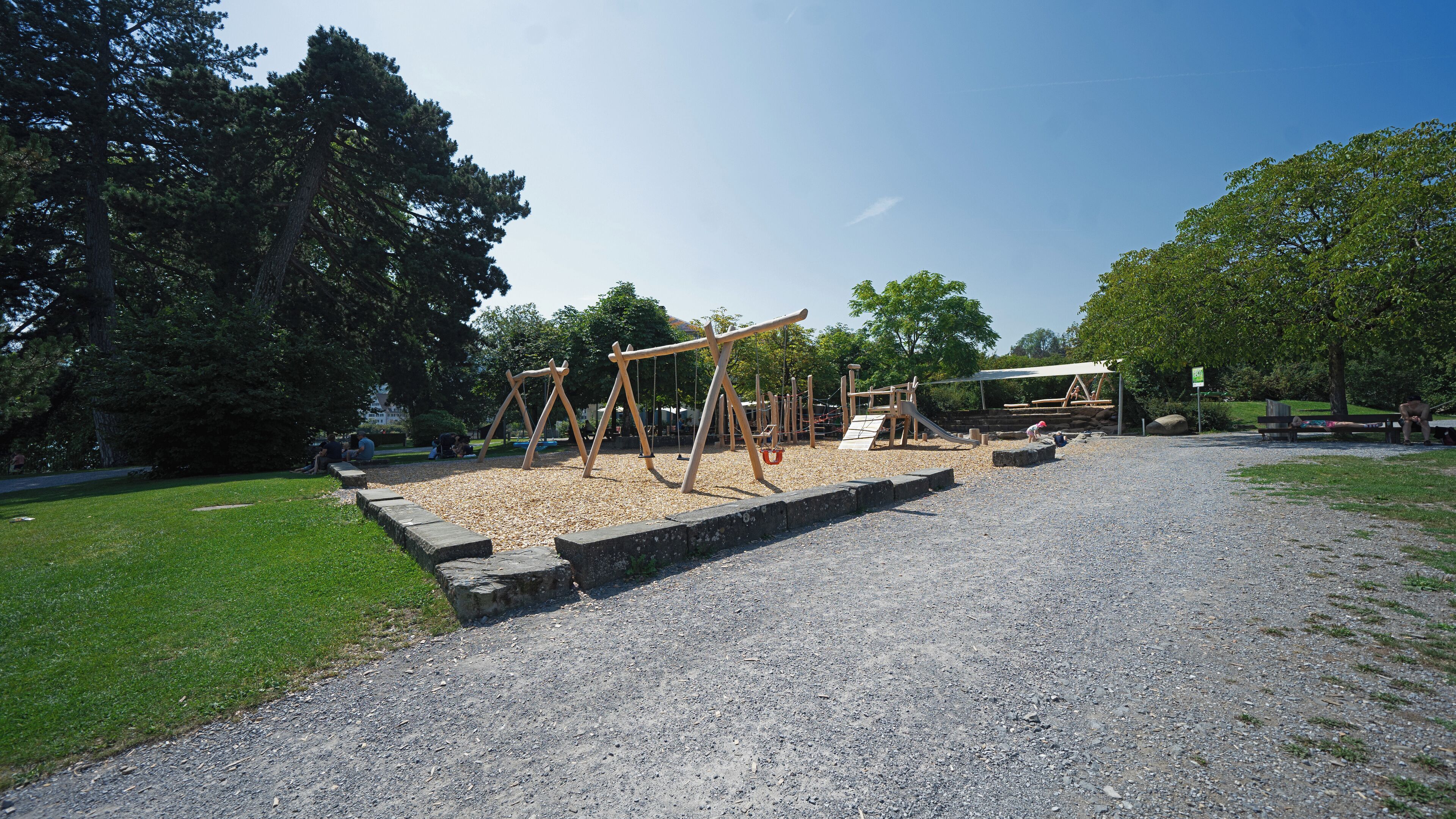 Un parque infantil con columpios y un tobogán, rodeado de árboles y un camino de grava.