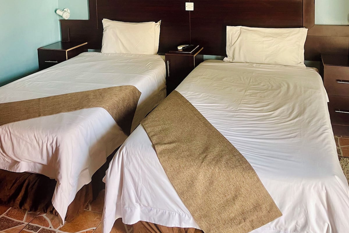 A hotel room with two single beds, wooden headboards, a nightstand with a lamp, and a tiled floor.