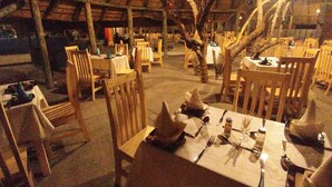 Un espace de restauration en plein air avec des tables et des chaises en bois, un toit de chaume et un arbre au centre.