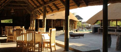 An outdoor dining area with wooden tables and chairs, thatched-roof structures, and a stone-paved floor.