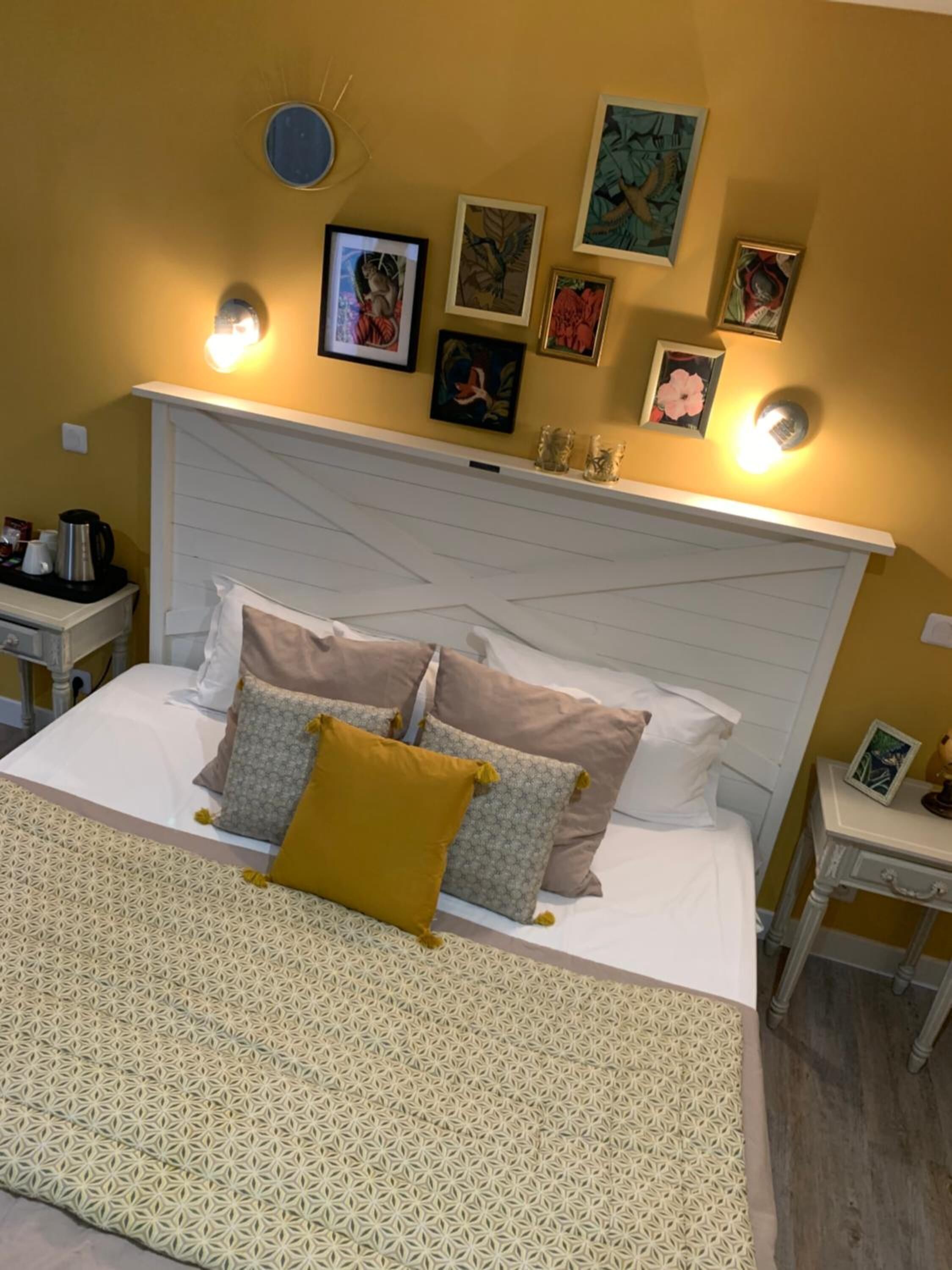 Ein ordentlich bezogenes Bett mit gemusterten Betttextilien und mehreren Kissen, ein Nachttisch mit Lampe sowie eine Wand mit gerahmten Kunstwerken.
