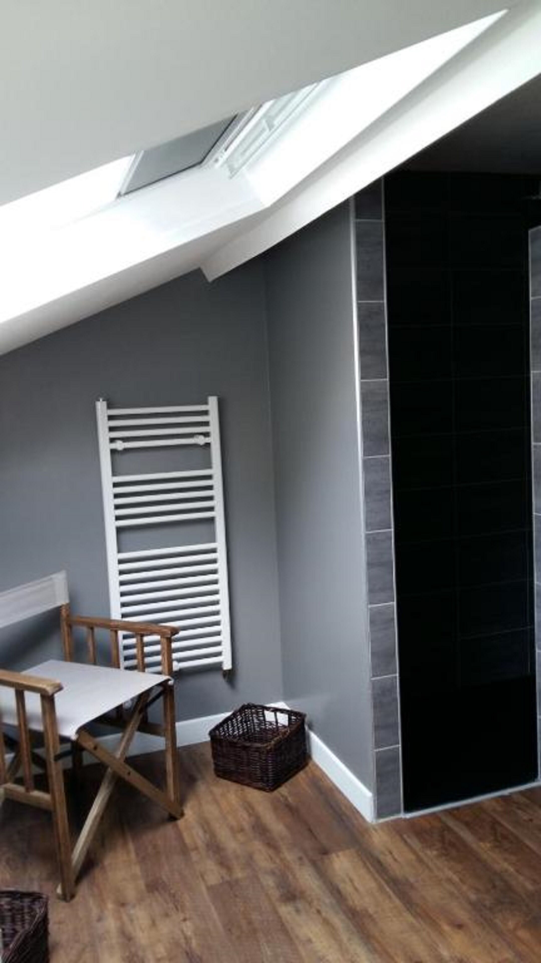Une pièce avec un plafond en pente, un radiateur blanc, une chaise en bois, un panier en osier et un mur recouvert de carreaux sombres.