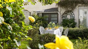 Un jardín con una mesa preparada para una comida, rodeado de follaje y un edificio al fondo.