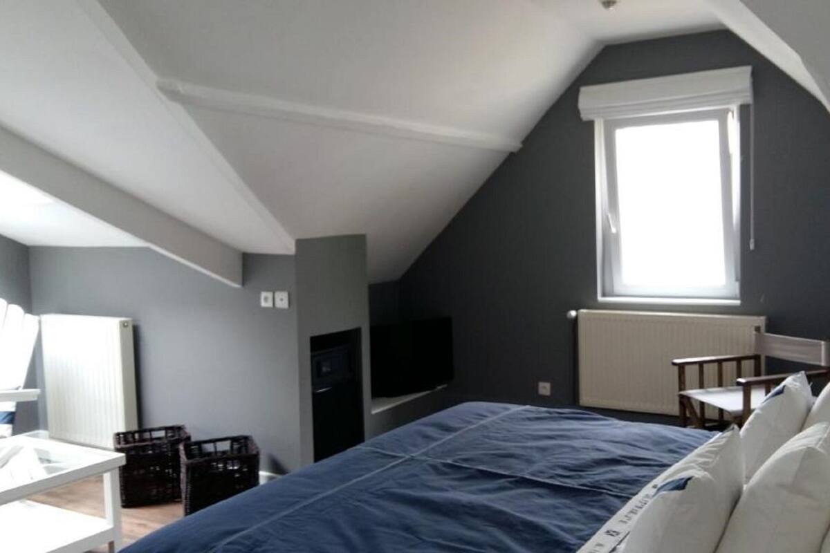 A bedroom with a sloped ceiling, a bed with white and blue bedding, a desk, a chair, a TV, and a window with a view.