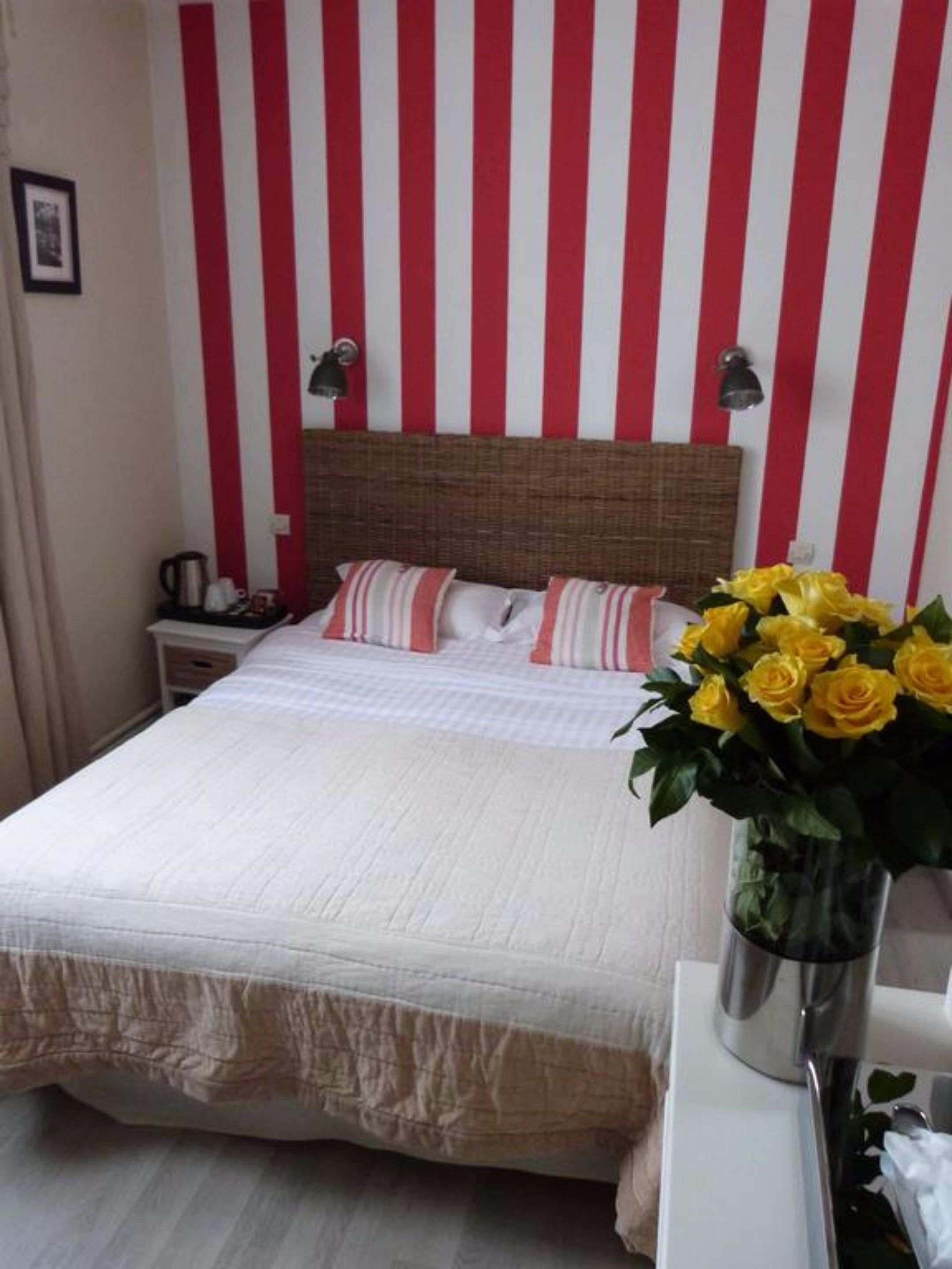 Een hotelkamer met een gestreepte rode en witte wand, een groot bed met witte beddengoed, twee bedlampen en een vaas met gele rozen.