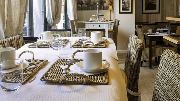 Un comedor con sillas de mimbre, una mesa larga dispuesta con platos, tazas y vasos, y lámparas colgantes arriba.