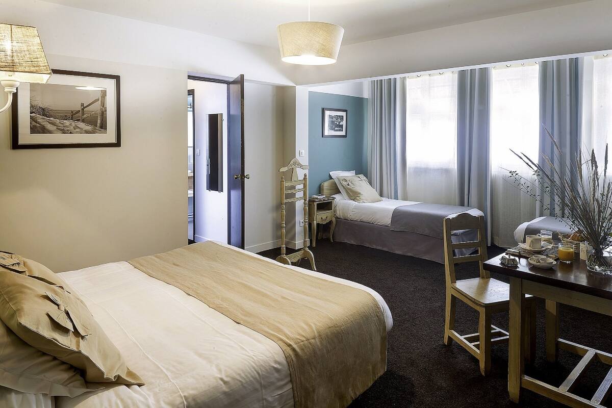 A hotel room with two beds, a dining table, and a chair. There is a picture on the wall and a lamp above the bed.