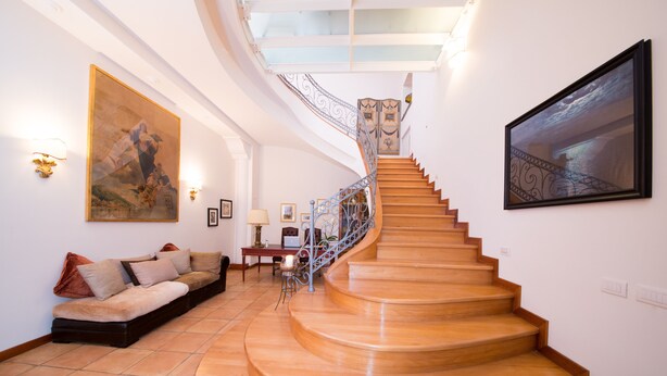 A grand staircase with a curved design, leading to a landing area with a sofa and a table. - B&B Maiurè (Maiori)
