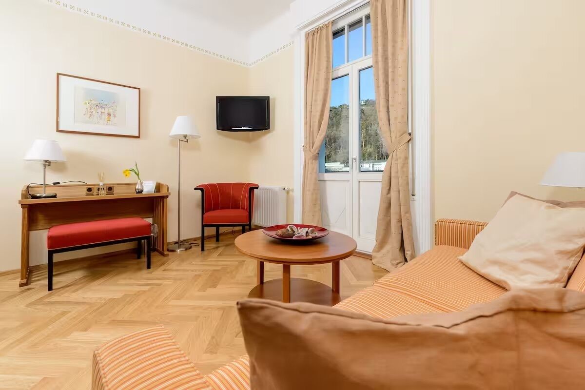 A hotel room with a wooden floor, a sofa, a red chair, a small table with a bowl, a TV mounted on the wall, and a window with curtains.