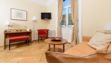 Habitación de hotel con suelo de madera, un sofá, una silla roja, una mesita con un bol, un televisor montado en la pared y una ventana con cortinas.